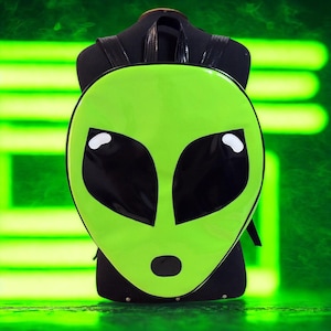 May include: A neon green backpack shaped like an alien face. The backpack features large black eyes, a small black nose, and white highlights. The straps and back of the backpack are black. The background is a blurred green.