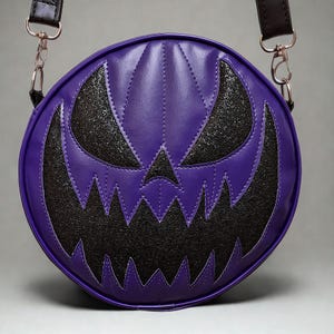 Pode incluir: Uma bolsa transversal redonda roxa com um design costurado. A bolsa apresenta um rosto de abóbora de Halloween preto e brilhante. Possui uma alça preta e ferragens prateadas. A bolsa é projetada para o Halloween.