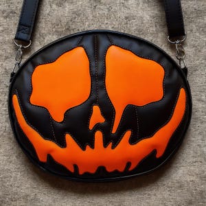 May include: Black and orange Halloween-themed crossbody bag. The bag is round with a stitched design of a jack-o'-lantern face. The bag has a black strap and silver-colored metal hardware. The pumpkin face is orange with black outlining.