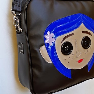 May include: Black shoulder bag featuring a cartoon face with blue hair, button eyes, and a flower. The bag has a silver zipper and a small metal tag that says "Nova Prado".