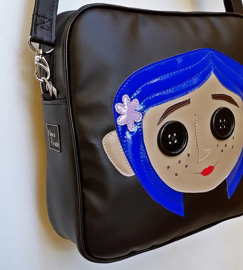 May include: Black shoulder bag featuring a cartoon face with blue hair, button eyes, and a flower. The bag has a silver zipper and a small metal tag that says "Nova Prado".