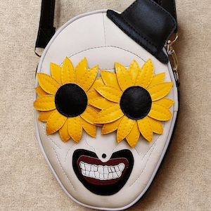 Peut inclure: Sac bandoulière blanc au design fantaisiste. Il présente un visage avec des yeux en tournesol, un chapeau haut-de-forme noir et une bouche souriante. Les tournesols sont jaunes avec des centres noirs. Le sac a une sangle noire.