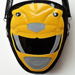 Pode incluir: Bolsa transversal preta e amarela com design de capacete Power Ranger. A bolsa tem um acabamento brilhante, com um rosto de capacete amarelo, detalhes cinza e fundo preto. Inclui uma alça preta e ferragens prateadas.