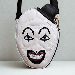 Può includere: Borsa a tracolla bianca e nera a forma di faccia da clown. La borsa presenta un cappello a cilindro nero, dettagli del viso cuciti e una chiusura con cerniera. Ha una tracolla nera e hardware color oro.