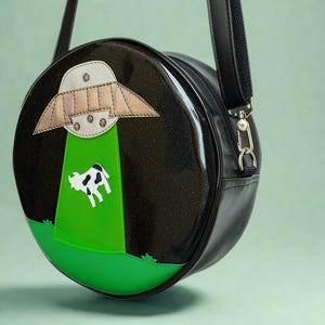 May include: A black, round crossbody bag with a whimsical design. The bag features an applique of a UFO beaming up a cow, set against a green field. The bag has a black strap and silver hardware.