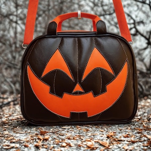 Pode incluir: Uma bolsa com tema de Halloween em forma de abóbora. A bolsa é principalmente marrom escuro com um rosto laranja sorridente. Possui uma alça e uma alça de ombro laranja. A bolsa está em uma superfície coberta de folhas.