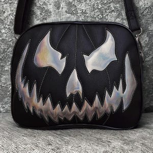 May include: Black crossbody bag with a jack-o'-lantern face design. The bag features a stitched outline and iridescent silver accents for the eyes, nose, and mouth. The bag has a black adjustable strap.
