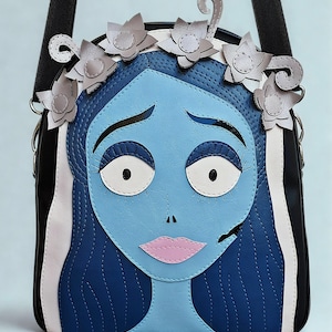 Pode incluir: Bolsa transversal azul e preta com uma ilustração estilizada de uma personagem com pele azul, olhos brancos grandes e lábios rosa. A bolsa tem uma coroa de flores e uma alça preta.