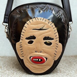 Pode incluir: Uma bolsa transversal marrom com um design de rosto costurado. A bolsa apresenta um rosto marrom claro com olhos pretos, boca vermelha e cabelo preto. A bolsa tem uma alça longa e ferragens de metal.