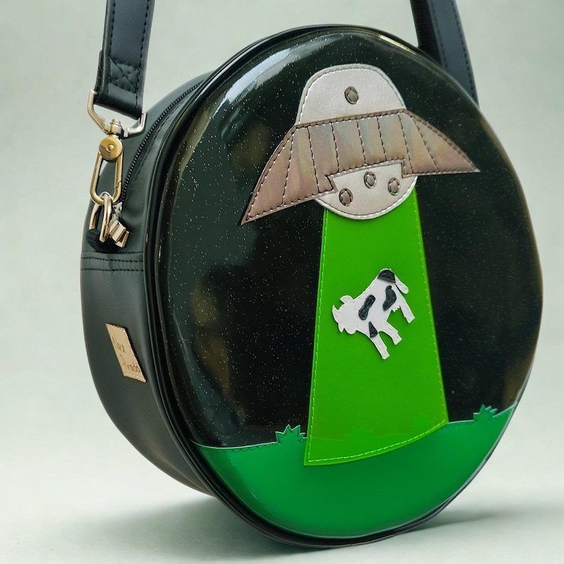 May include: A black, round handbag with a shiny finish. The design features a silver and brown UFO beaming a green light onto a black and white cow. The bag has a black strap and gold hardware.