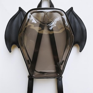 Puede incluir: Una mochila hexagonal transparente con correas negras y alas de murciélago. La mochila tiene un asa superior y correas ajustables para los hombros. Las alas son negras y están hechas de un material texturizado. Cuelga sobre un fondo blanco.
