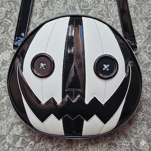 Pode incluir: Uma bolsa tiracolo redonda com um design de rosto de abóbora em preto e branco brilhante. A bolsa tem dois grandes olhos de botão pretos com costura branca, um nariz triangular e uma boca serrilhada. Inclui uma alça de ombro preta.