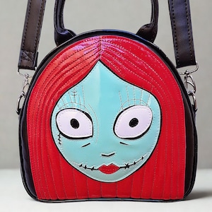 May include: A round handbag with a cartoon face design. It features red hair, a light blue face, and stitched details. The bag has a black body, a top handle, and a detachable shoulder strap. The design is inspired by a character from an animated film.