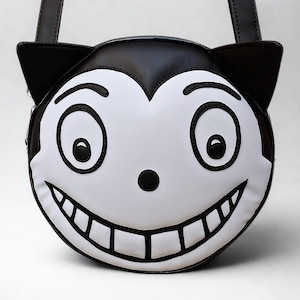 Pode incluir: Uma bolsa redonda em preto e branco com um design de rosto de gato de desenho animado. A bolsa apresenta um rosto sorridente com olhos grandes, um nariz e um sorriso largo. A bolsa tem uma alça preta e orelhas de gato.