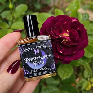 May include: A small rectangular glass bottle of perfume oil with a black cap. The label features a purple crescent moon, a butterfly, and the text Violet Apothic, Persephone's Path, Perfume Oil, and Net 1/2 oz / 15 ml in white lettering.