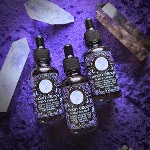 May include: Three glass bottles with dropper tops filled with a dark liquid. Each bottle has a black and white label with the text "Moon Drops Herbal Tincture" and a moon illustration. The text on the label also reads "A potent sleep-inducing blend to support deep rest and vivid dreaming. 1 oz. Violet Apothic."