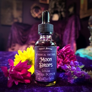 May include: A glass bottle of "Moon Drops" botanical tincture with a black dropper. The label reads "Violet Apothic" and "Lunar Dream Potion." The bottle is surrounded by purple and yellow flowers, set against a purple velvet surface.