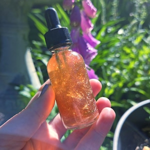 May include: A small, clear glass bottle with a black dropper filled with a shimmering, golden-colored liquid. The bottle is held in front of a blurred background of green foliage and purple flowers, suggesting a natural setting.