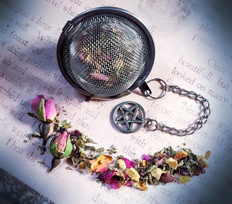 May include: A silver tea infuser with a pentagram charm on the chain, filled with loose leaf tea and rose buds. The infuser is sitting on a piece of paper with text.