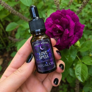 May include: A small amber glass dropper bottle with a black cap and a rectangular label. The label features a purple and blue cosmic background with a crescent moon, white text reading VIOLET APOTHIC, LOVE SPELL, and RITUAL OIL.