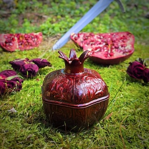 May include: A decorative, burgundy-colored ceramic pomegranate-shaped container with a lid, set on a bed of green moss. The container has an embossed design. Slices of red pomegranate and dried rose buds are scattered around the container.