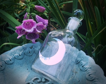 Könnte beinhalten: Eine klare Glasflasche mit einem Halbmond-Design auf der Vorderseite. Die Flasche steht auf einer blauen und weißen Keramikfläche.