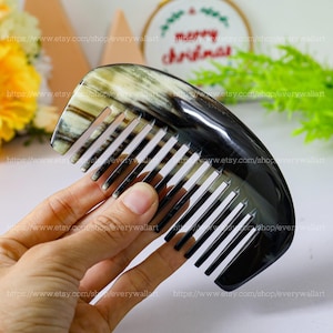 Puede incluir: Un peine curvo con una mezcla de tonos negros, blancos y beige. Los dientes del peine están espaciados uniformemente y parecen estar hechos de un material de color claro. El peine se sostiene en una mano.