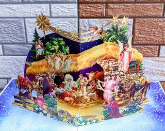 Può includere: Un biglietto di Natale pop-up che rappresenta una scena della Natività con una stalla, una mangiatoia, un cammello, un asino e figure di Maria, Giuseppe, il bambino Gesù e angeli. Il biglietto è decorato con stelle dorate e un cielo stellato blu e bianco.
