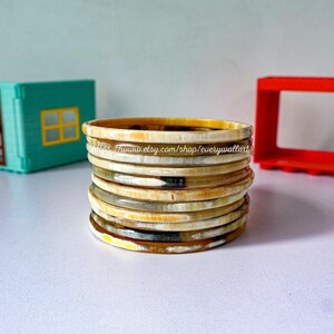 May include: A stack of 10 horn bangles in shades of cream, brown, and black. The bangles are stacked on a white surface.