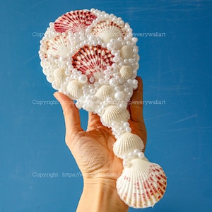 May include: A hand mirror decorated with white and pink seashells and white pearls. The mirror is shaped like a seashell and has a long handle.