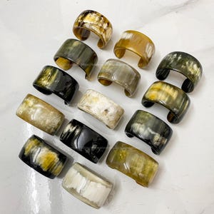 May include: A collection of 14 horn bracelets in various colours and patterns, including black, white, brown, and yellow. The bracelets are made of natural horn and have a polished finish.