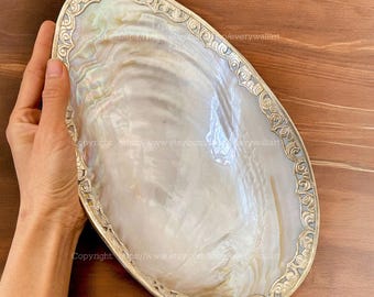 May include: An oval-shaped, iridescent pearl shell tray with a decorative silver-colored border. The tray is held by a hand, showcasing its size and natural beauty. The shell's surface reflects light, displaying a range of colors.
