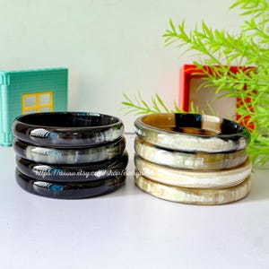 May include: A set of four black and white horn bangles and a set of four natural horn bangles. The bangles are stacked on top of each other.
