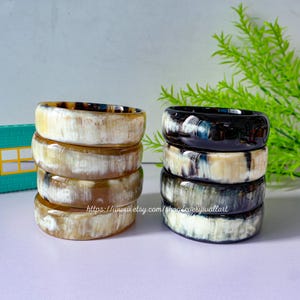 May include: Two stacks of horn bangles, one stack is black and white, the other is brown and white. The stacks are on a white surface with a green and white background.