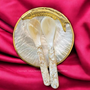 Könnte beinhalten: Zwei weiße Perlmuttlöffel mit goldenen Flocken in den Griffen, die auf einem weißen Perlmuttteller mit einem goldenen Fischdesign liegen.