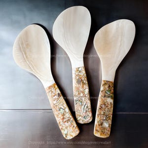 May include: Three white wooden serving spoons with iridescent mother-of-pearl handles. The handles are made of a mixture of small pieces of seashells in shades of white, brown, and green.