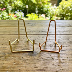 May include: Two metal display stands, one gold-toned and one copper-toned, are positioned on a wooden surface. The stands have a simple, minimalist design with a triangular shape and small, curved supports to hold items.