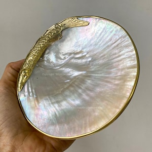 Può includere: Un piatto decorativo a forma di conchiglia bianca iridescente con un accento di pesce color oro. Il piatto ha un bordo color oro e una finitura perlata. Il pesce è dettagliato con squame e pinne.