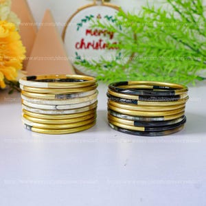 Può includere: Un set di 10 braccialetti in corno dorati e neri. I braccialetti sono impilati su una superficie bianca.