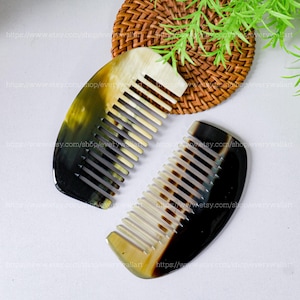 May include: Two handcrafted horn combs. The combs have a curved shape with closely spaced teeth. The colors are a mix of black, brown, and cream. The combs are displayed on a white surface with a woven coaster and greenery in the background.