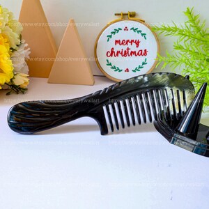 Puede incluir: Un peine de cuerno negro y blanco con un mango decorativo. El peine está sobre una superficie blanca junto a un pequeño aro bordado blanco con el texto "merry christmas" en rojo.