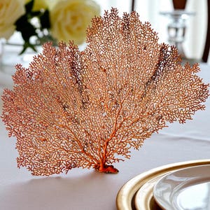 May include: An ornamental coral-like piece in orange and brown hues, shaped like a fan. The detailed design showcases a complex network of fine branches, giving it a natural, organic appearance. It rests on a white tablecloth, close to a gold-edged plate, suggesting a table setting.