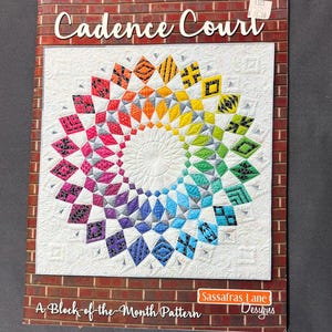 May include: A book cover featuring a colorful quilt design. The quilt has a circular pattern with a gradient of colors, including red, orange, yellow, green, blue, and purple. The title "Cadence Court" is displayed at the top, with "A Block-of-the-Month Pattern" at the bottom.