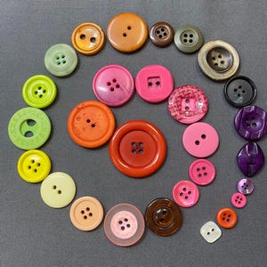 May include: A collection of colorful buttons in various sizes and shapes, including round, square, and oval. The buttons are in shades of green, yellow, orange, pink, brown, and purple. Some buttons have two or four holes, while others have a shank.