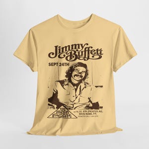 Może przedstawiać: Żółta koszulka z brązowym nadrukiem Jimmy'ego Buffetta. Nadruk zawiera portret Jimmy'ego Buffetta i tekst "Jimmy Buffett" w stylizowanym kroju pisma. Tekst "Sept 24th" jest nadrukowany pod nazwiskiem. Tekst "The Main Point" jest nadrukowany w kształcie trójkąta pod portretem. Tekst "On Rt. 30, 874 Lancaster Ave. Bryn Mawr, PA. Tickets Call LAS-3375" jest nadrukowany pod trójkątem.