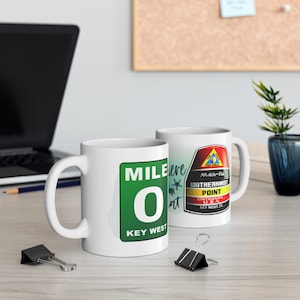 Puede incluir: Dos tazas de cerámica blanca. Una taza presenta un letrero verde con "MILE 0 KEY WEST" en texto blanco. La otra taza muestra un diseño colorido con "SOUTHERNMOST POINT CONTINENTAL U.S.A." y "KEY WEST, FL."