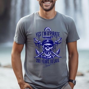 Pode incluir: Camiseta cinza com um gráfico azul de uma caveira e ossos cruzados com uma âncora. O texto "Yes I'm a Pirate" e "200 Years to Late" está impresso na camiseta.