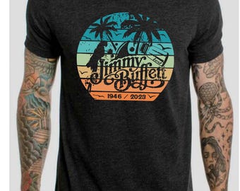Op de afbeelding: Een donkergrijs T-shirt met een vintage-stijl grafische afbeelding van palmbomen, een gitaar en de tekst "Jimmy Buffett 1946 / 2023".