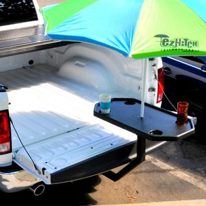 May include: A truck bed accessory featuring a black table with cup holders and a multi-coloured umbrella. The umbrella is blue and green. The table is attached to the truck bed and holds two drinks.