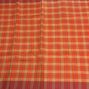 May include: A close-up of a plaid fabric with a repeating pattern of orange, tan, and red squares. The fabric has a textured appearance and is likely used for clothing or home decor.